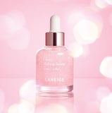 A bottle of Laneige Glowy Makeup Serum, with a rose-gold cap and dropper, sits on a reflective surface against a soft pink background, promising a radiant and healthy glow for long-lasting makeup.
